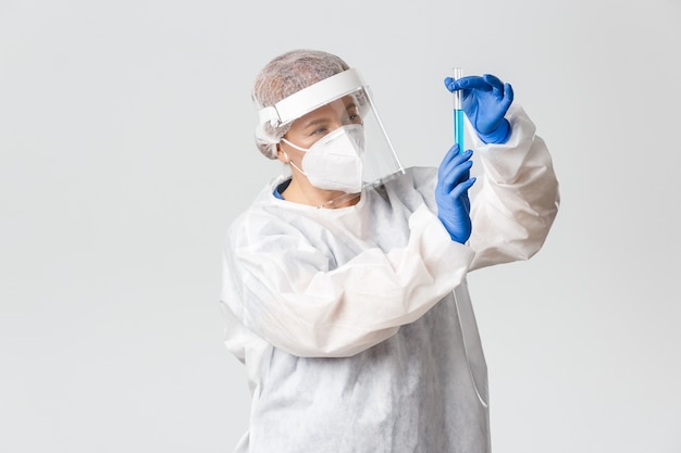 Personnel médical, pandémie de covid-19, concept de coronavirus. Femme médecin réfléchie et professionnelle en équipement de protection individuelle pensant tout en regardant tube à essai avec vaccin, fond gris.