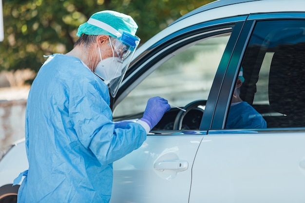 Personnel médical portant un EPI, effectuant une PCR avec un écouvillon à la main, sur un patient à l'intérieur de sa voiture pour détecter s'il est infecté par le COVID-19