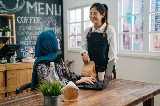 personnel de serveuse de fille asiatique en tablier tenant une tasse de café jetable et un sac en papier dans un café-bar donnant à une cliente musulmane sur le siège. travailleuse malaise indépendante travaillant sur un ordinateur portable souriant pour partir.