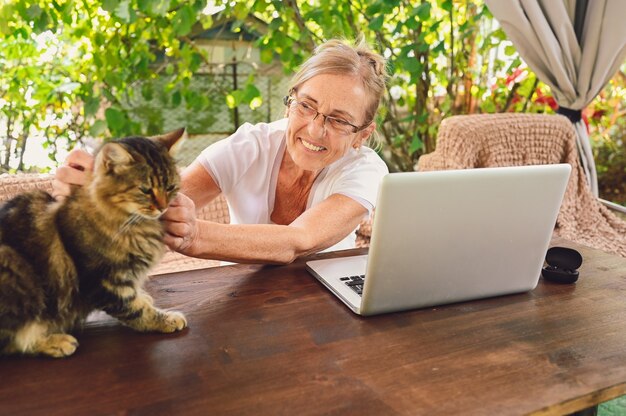 personnes âgées happy senior woman avec chat domestique utilisent des écouteurs sans fil travaillant en ligne avec un ordinateur portable en plein air dans le jardin. Travail à distance