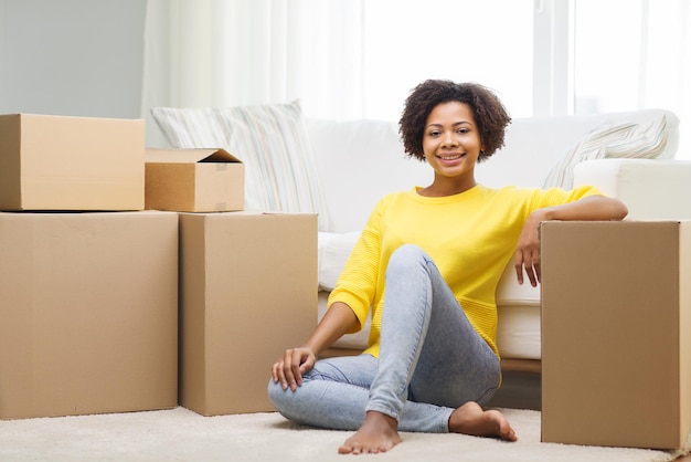 personnes, déplacement d'un nouvel endroit et concept de réparation - jeune femme afro-américaine heureuse avec de nombreuses boîtes en carton assises sur le sol à la maison