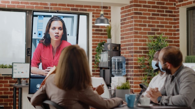 Les personnes avec un masque facial parlent par téléconférence à une femme, en utilisant la vidéoconférence en ligne sur le moniteur. Collègues faisant des télécommunications par vidéoconférence à distance pendant la pandémie.