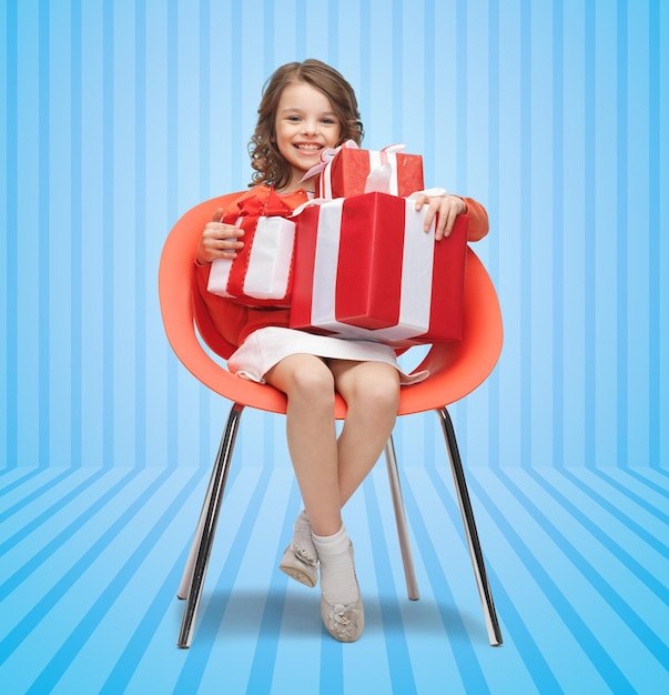 personnes, noël, vacances, cadeaux et concept d'enfance - petite fille heureuse avec des coffrets cadeaux assis sur une chaise sur fond rayé bleu