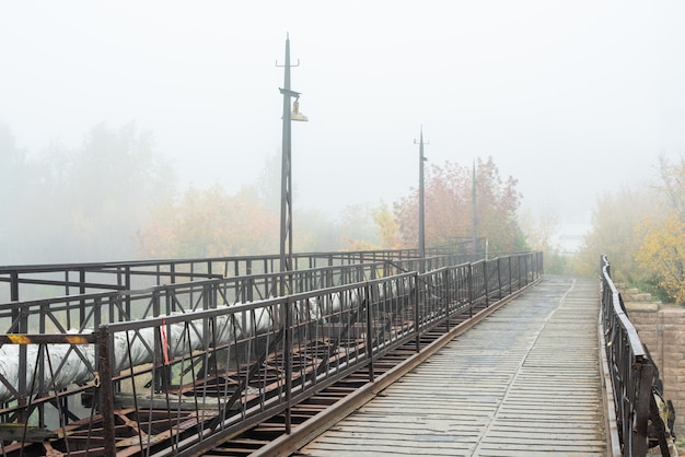 Perspective d'une longue route ou d'un pont entouré d'une clôture métallique, d'un tuyau et d'arbres au feuillage jaune le matin d'automne boudeur et brumeux
