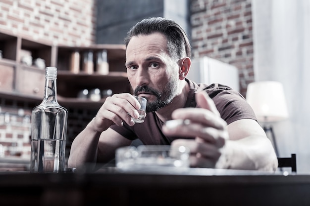 À perte. Déprimé triste homme sans joie mettant sa main sur la table et buvant de la vodka sans savoir quoi faire à propos de son problème