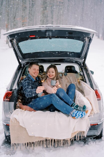 Un petit ami et une petite amie souriants sont assis dans le coffre d'une voiture sous la neige avec des étincelles et
