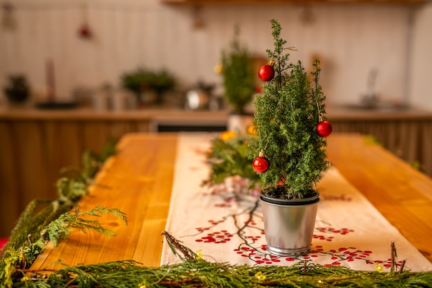 Un petit arbre de Noël avec des boules rouges dans un seau en métal se dresse sur une table en bois dans la cuisine décorée.