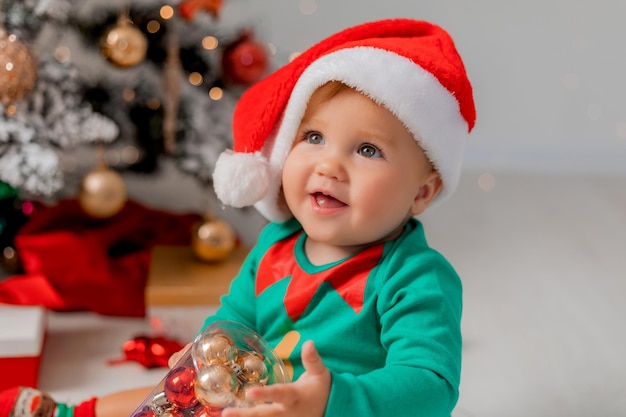 Le petit assistant du Père Noël à côté du sapin de Noël avec des boules de Noël dans ses mains. portrait d'un bébé. enfant en costume d'elfe. concept du nouvel an d'hiver. espace pour le texte. Photo de haute qualité