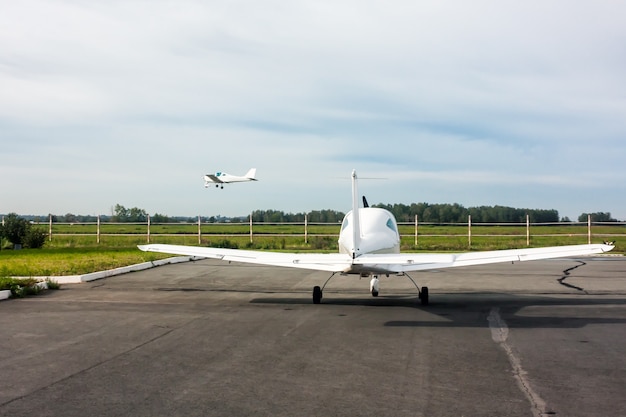 Un petit avion de sport se tient sur le parking de l'aérodrome et l'autre avion arrive pour atterrir