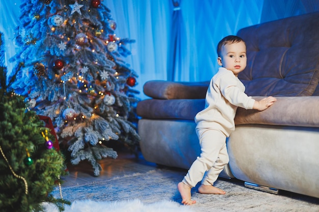 Petit beau garçon d'un an vêtu d'un costume beige sur fond d'arbre de Noël dans un environnement familial Les émotions des enfants avant de célébrer les vacances du Nouvel An