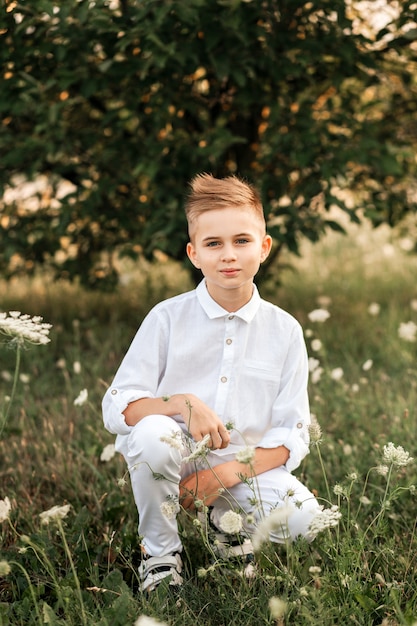 Un petit beau garçon blond dans une chemise blanche et un pantalon blanc dans la nature en été. Enfance heureuse. Émotions positives