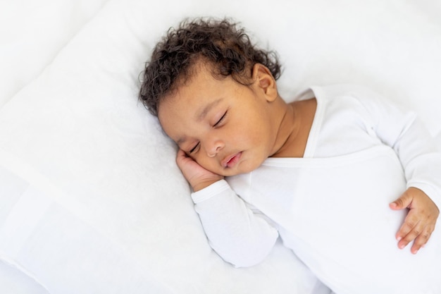 Un petit bébé afro-américain dort sur un lit blanc à la maison avec sa main repliée sous sa joue