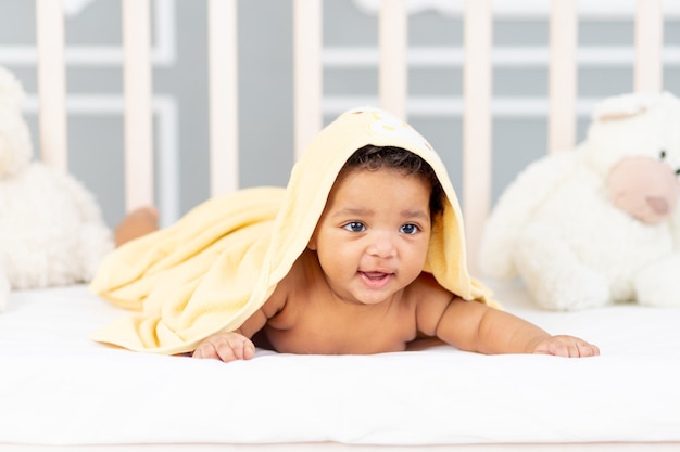 Un petit bébé afro-américain souriant est allongé dans son lit après un bain dans une serviette jaune