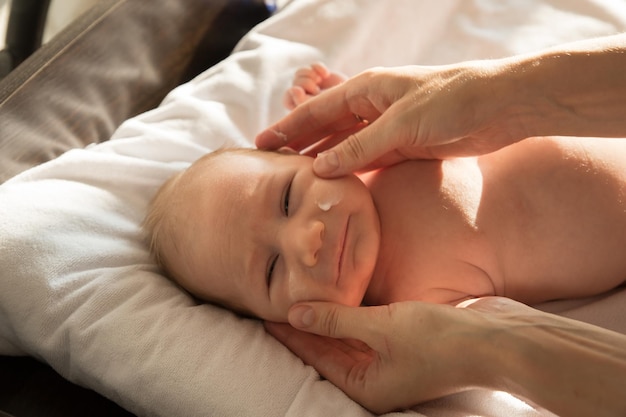 Un petit bébé allongé sur un lit sur des draps blancs pendant que son parent applique de la crème sur son visage