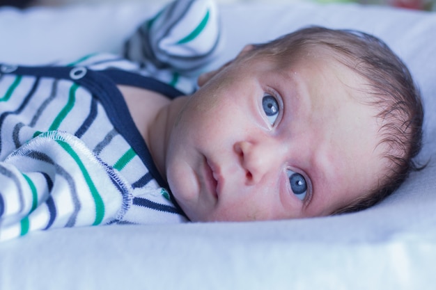 Un petit bébé aux yeux bleus dans un costume rayé est allongé sur un oreiller blanc et regarde légèrement sur le côté ...