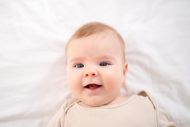 Un petit bébé en bonne santé dans un body en coton est allongé sur le dos sur un lit sur une literie blanche dans la chambre à la maison regarde la caméra sourit un grand portrait de l'enfant