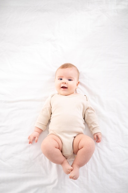 Un petit bébé en bonne santé dans un body en coton est allongé sur le dos sur un lit sur une literie blanche dans la chambre à la maison regarde la caméra sourit