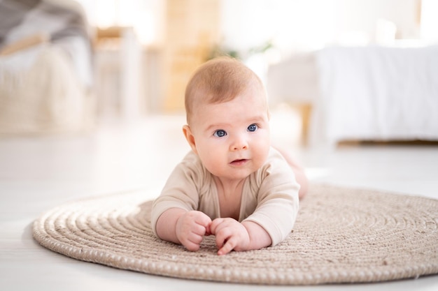 Un petit bébé en bonne santé dans un body en coton est allongé sur le ventre sur un tapis dans le salon lumineux de la maison regarde la caméra sourit