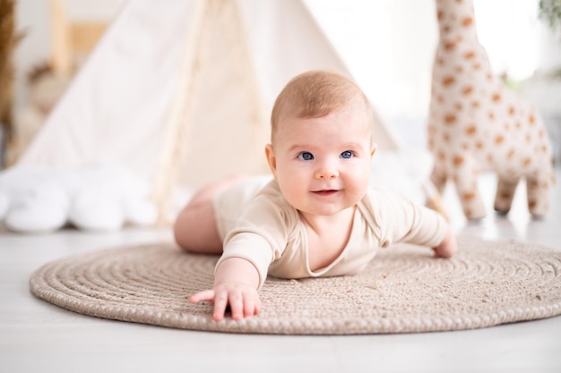 Un petit bébé en bonne santé dans un body en coton est allongé sur le ventre sur un tapis dans le salon de la maison sur fond de wigwam et de jouets en peluche