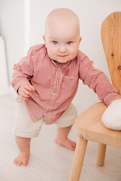 Petit bébé caucasien en chemise rouge, short blanc se tient près de la chaise en bois