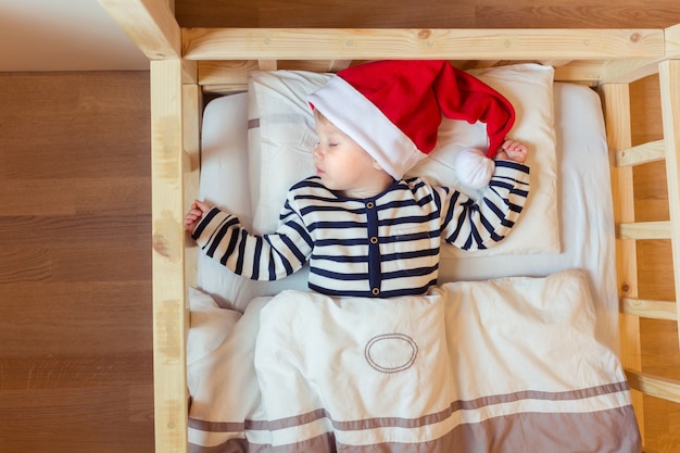 Un petit bébé dort sur un lit blanc dans un chapeau de père Noël rouge avant la nuit de Noël
