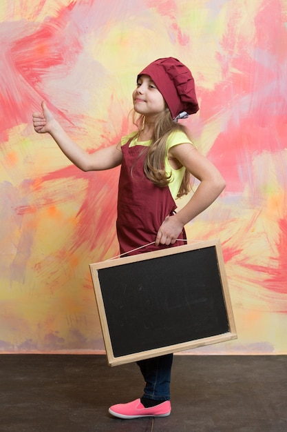 Petit bébé fille ou enfant mignon avec un visage heureux en toque rouge et tablier de cuisinier tenant un tableau noir montrant le pouce vers le haut sur fond abstrait coloré cuisine copie espace