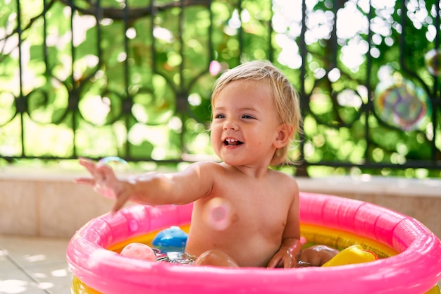 Un petit bébé souriant est assis dans une petite piscine gonflable et attrape des bulles de savon