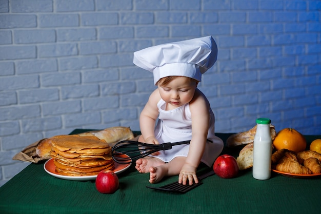 Petit bel enfant vêtu d'un tablier, costume de cuisine pour enfants. Crêpes et pâtisseries fraîches sur la table