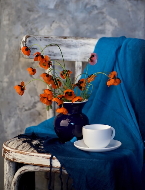 Un petit bouquet de coquelicots rouges dans un vase bleu sur une chaise vintage. Coquelicots et tasse de café.