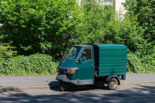 Un petit camion de livraison vert est garé sur le trottoir à côté d'une rangée d'arbres