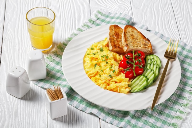 Petit-déjeuner américain composé d'œufs brouillés avec du concombre tranché, des tomates, du pain grillé et un verre de jus d'orange frais sur une plaque blanche sur une table texturée en bois