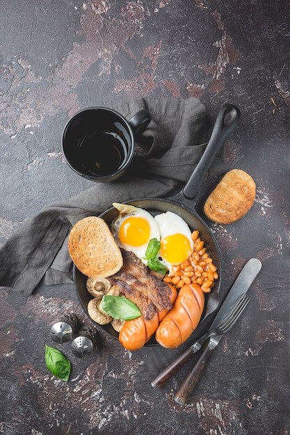 Petit-déjeuner anglais complet avec œufs au plat, saucisses, bacon, haricots, pain grillé et tomates sur pierre sombre