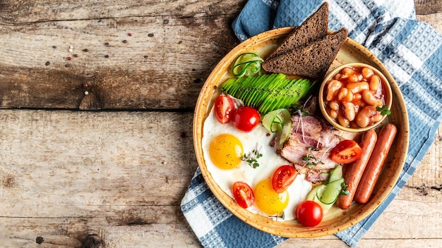 Petit-déjeuner anglais complet avec œufs frits, bacon, haricots, toasts, tomates, saucisses et avocat sur fond de bois, vue de dessus, bannière de frais généraux