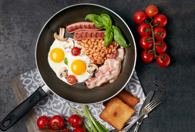 Petit-déjeuner anglais dans une casserole avec des œufs au plat, des saucisses, des haricots, des tomates, des champignons, du bacon et du pain grillé sur une table en bois. Copiez l'espace. Vue de dessus