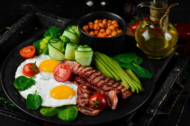 Petit-déjeuner anglais : pois chiches, avocats, œufs brouillés, légumes au bacon sur une assiette en pierre noire. Vue de dessus. Espace libre pour le texte.