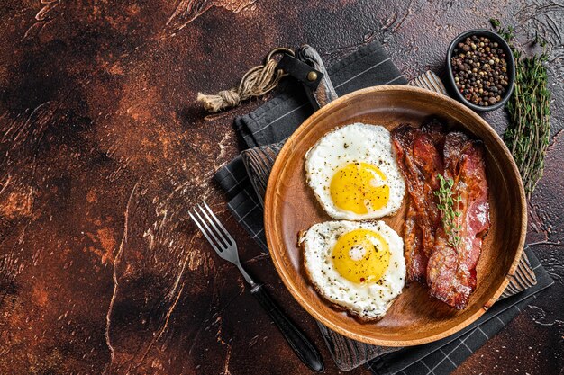 Petit-déjeuner anglais traditionnel avec œufs au plat et bacon dans une assiette en bois. Fond sombre. Vue de dessus. Espace de copie.