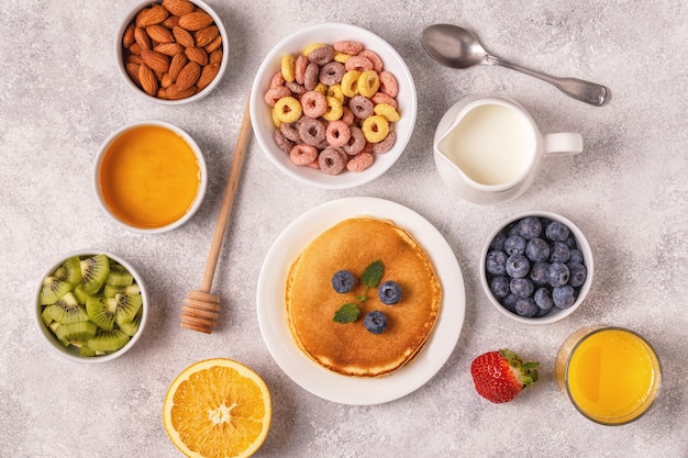 Petit-déjeuner avec anneaux de céréales colorés, crêpes, fruits, lait, jus