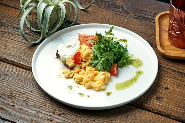 Petit-déjeuner appétissant - œufs brouillés, avec roquette, saumon et pain grillé avec du yaourt grec dans une assiette sur une table en bois