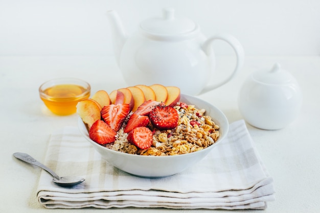 Photo petit-déjeuner bouillie d'avoine avec céréales granola fraises pêches miel dans une assiette blanche sur le fond d'une théière blanche et bols avec du miel sur une assiette blanche
