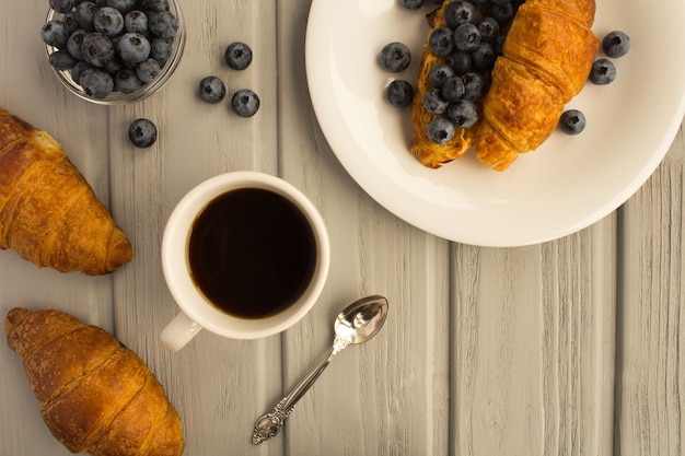 Petit déjeuner : café, croissant avec pâte d'arachide et bleuets sur le fond en bois gris. Vue de dessus. Espace de copie.