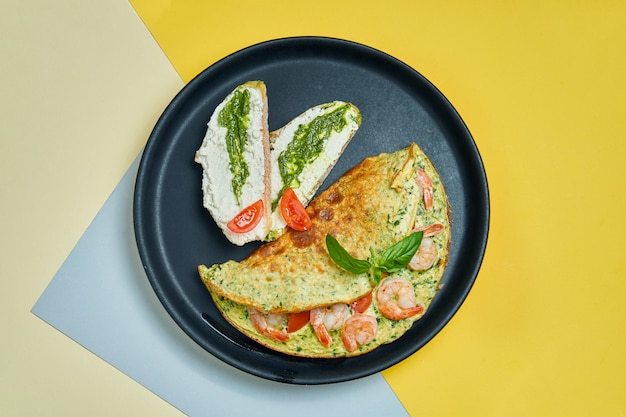 Petit déjeuner continental - omelette aux crevettes, tomates et basilic avec du pain grillé avec du fromage à la crème et du pesto sur une plaque noire sur s.