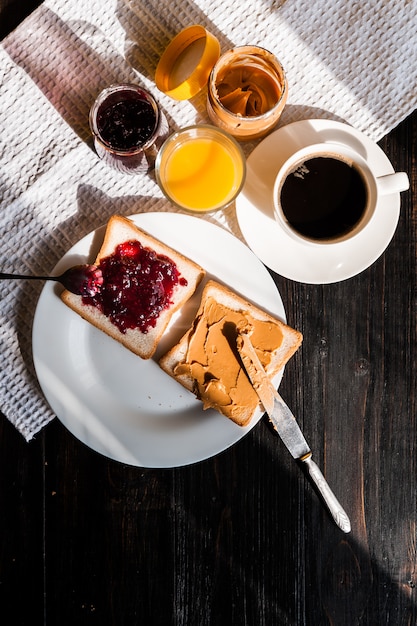 Petit déjeuner avec du beurre de cacahuète et de la confiture et une tasse de café sur une table en bois au soleil du matin