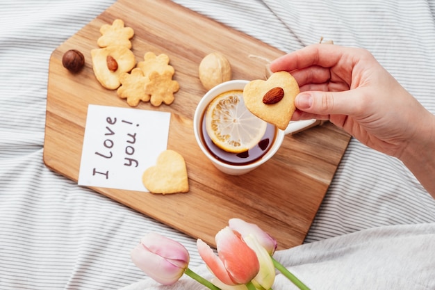 Petit Déjeuner Festif Au Lit Pour La Saint-valentin. Thé Et Biscuits De Vos Propres Mains En Forme De Cœur. Une Note Sur Papier, Un Cadeau Et Des Fleurs à Votre Fille Bien-aimée.