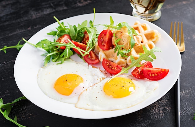 Petit-déjeuner avec gaufres au fromage, œufs au plat, tomate, houmous et roquette sur table sombre. Apéritifs, goûter, brunch. Nourriture végétarienne saine.