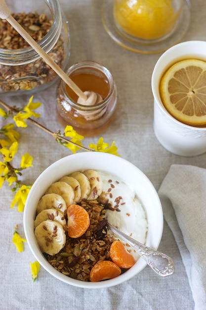 Petit déjeuner de granola avec du yaourt et du thé et des fleurs de forsythia sur une nappe en lin. Style rustique.