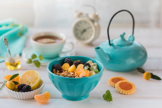 Petit déjeuner avec granola, fruits, jus fraîchement pressé et thé sur une table blanche.