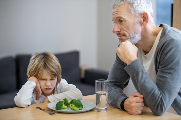 Petit-déjeuner. Homme mûr essayant de faire manger sainement à son fils tandis que le garçon a l'air malheureux