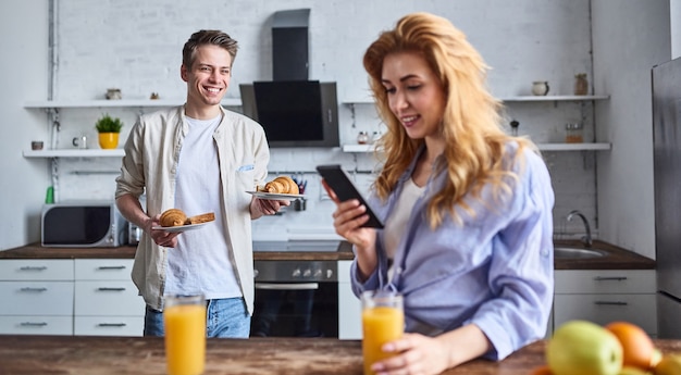 Petit-déjeuner d'un jeune couple amoureux à la cuisine à domicile. Belle fille avec un téléphone à la main boit du jus. Petit-déjeuner préparé par petit ami.