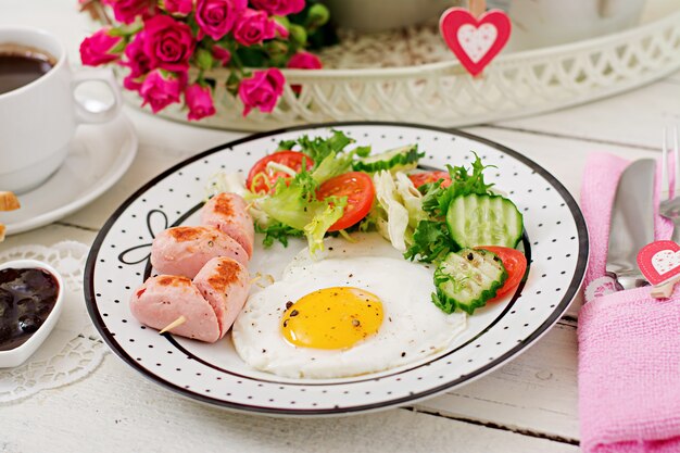 Petit déjeuner le jour de la Saint-Valentin - œuf au plat en forme de cœur, toasts, saucisses et légumes frais. Tasse de café. Petit déjeuner anglais