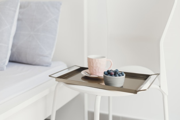 Petit-déjeuner léger servi sur un plateau en métal. Café dans une tasse rose et myrtille fraîche sur une chaise blanche